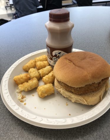  A plate from the Oakdale High Cafeteria containing a chicken sandwich, tater tots, and chocolate milk