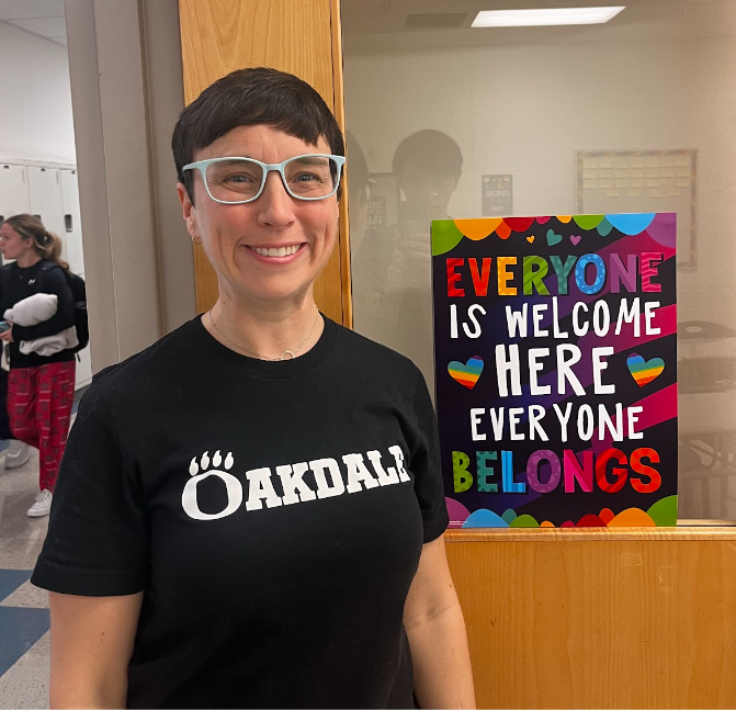 Ms. Sherman, in her Oakdale spirit wear, poses with a sign on her classroom door that reads, “Everyone is welcome here, everyone belongs.”